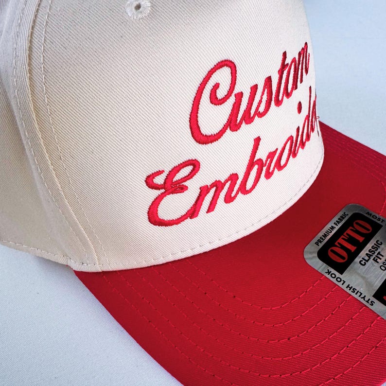 May include: A cream-colored baseball cap with a red bill and the words "Custom Embroidery" embroidered in red script. The cap has a classic fit and a stylish look.
