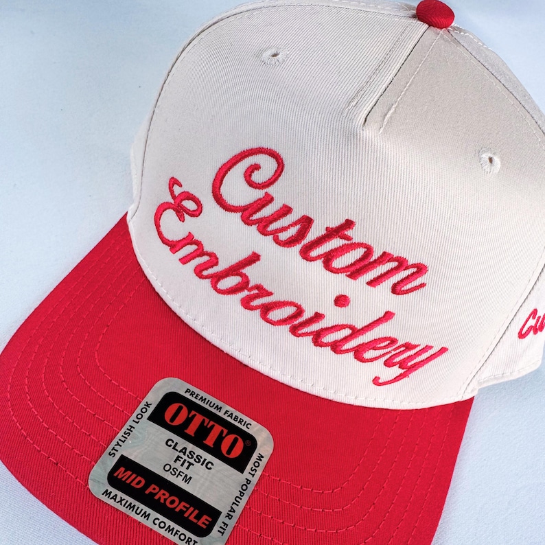 May include: A baseball cap with a red bill and button, and a cream-colored crown. The words "Custom Embroidery" are embroidered in red script across the front. A sticker on the bill reads "OTTO" and "Mid Profile".