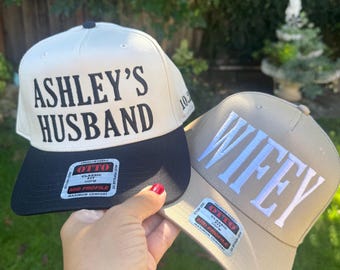 May include: Two baseball caps, one cream and black, the other tan. The cream cap reads "ASHLEY'S HUSBAND" in black, the tan cap reads "WIFEY" in white. Both have "OTTO" tags.
