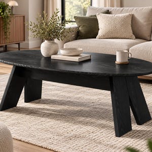 May include: An oval coffee table with a black wood grain finish, featuring a smooth tabletop and four angled, thick slab legs. The table is topped with a light-coloured ceramic vase containing greenery, a shallow bowl, a book, and a ribbed mug.
