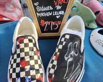 May include: A pair of black and white checkered Vans slip-on shoes with red blood splatter. One shoe features a black and white illustration of the Ghostface killer from the Scream movie franchise. The shoes are signed by Matthew Lillard, who played Stu Macher in the first Scream film. The sign reads "Scream Vans Signed by Matthew Lillard $275".