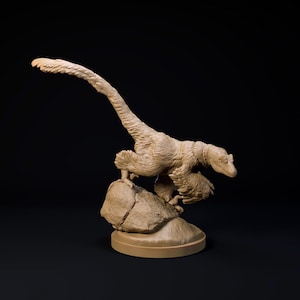 May include: A tan-coloured dinosaur figurine, a raptor, is perched atop a rock. The dinosaur has detailed feather-like textures and is posed with its tail extended. The base is a circular platform, and the background is black.