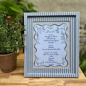 May include: A framed menu with a blue and white striped border. The menu lists drink options including wine, beer, and spirits. The text is in a decorative font with floral accents. The frame is on a wooden surface.