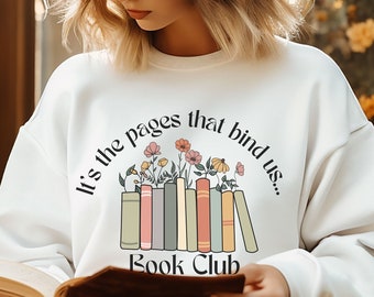 May include: White sweatshirt with the text "It's the pages that bind us... Book Club" above a graphic of books and flowers. The person in the image is wearing glasses and reading a book. The background includes a window and yellow flowers.