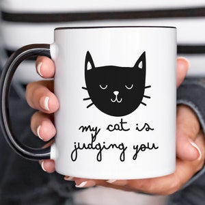 May include: White ceramic mug with a black handle. The mug has a black and white illustration of a cat with the text "my cat is judging you".