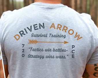 Könnte beinhalten: Ein graues T-Shirt mit einem braunen und blauen Logo, auf dem "Driven Arrow Survival Training" steht und der Text "Tactics win battles - Strategy wins wars" enthalten ist.