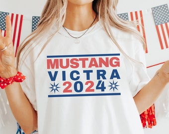 Könnte beinhalten: Weißes T-Shirt mit rot-weiß-blauem Text, der "Mustang Victra 2024" lautet, mit einem Stern auf jeder Seite des Jahres. Die Person, die das Shirt trägt, hält ein Glas mit roter Flüssigkeit und einer Erdbeere darin.