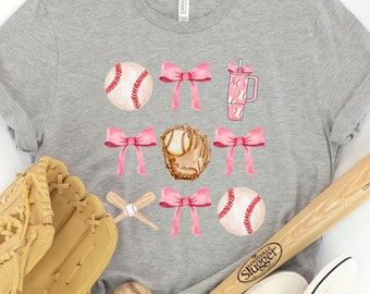 May include: A gray t-shirt with a baseball theme featuring pink bows, baseballs, a baseball glove, and a baseball bat. The shirt is surrounded by baseball equipment, including a baseball glove, a baseball bat, and baseballs.
