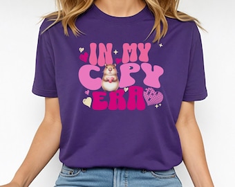 May include: A purple t-shirt with the text "IN MY CAPY ERA" in pink and white bubble letters. A cartoon capybara holding a heart is in the center. Pink hearts and stars are scattered around the text.