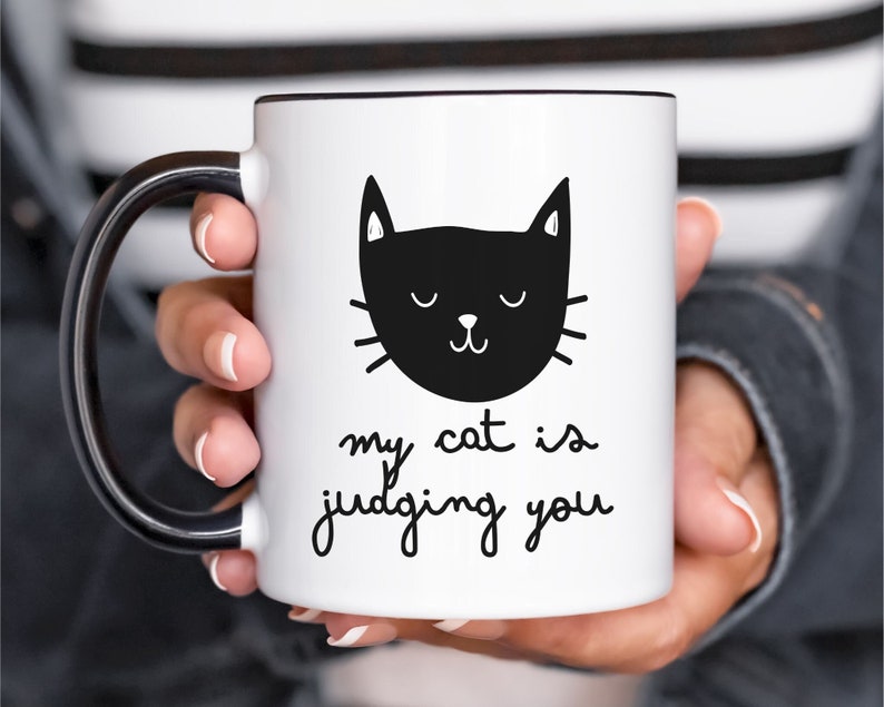 May include: White ceramic mug with a black handle. The mug has a black and white illustration of a cat with the text "my cat is judging you".