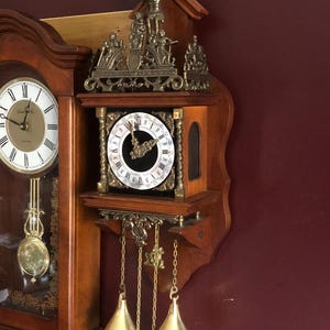 May include: A wooden wall clock with two clock faces. The clock features a pendulum, gold-coloured weights, and decorative metal accents. The top of the clock has a statue of a person holding a blue sphere.