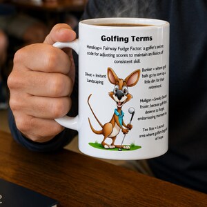 May include: A white ceramic mug featuring a cartoon kangaroo holding a golf club and definitions of Golfing Terms including Handicap, Divot, Bunker, Mulligan, and Tee Box printed in black text on the side of the mug with steam rising from the top.