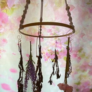 May include: A white macrame hanging mobile with a wooden ring and dried flowers and herbs hanging from it. The mobile is hanging from a white ceiling with a floral pattern.