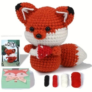 May include: A crocheted fox toy with orange, white, and black details. The fox holds a red and white mushroom. The image includes a DIY kit, a fox-themed gift box, and spools of yarn in black, red, and white.