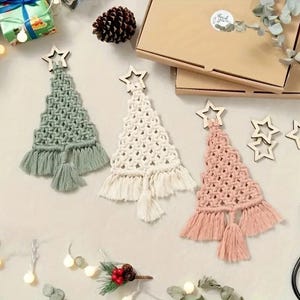 Pode incluir: Três enfeites de árvore de Natal em macramé verde sálvia, branco e rosa blush, cada um encimado por uma estrela de madeira. As árvores têm uma base com franjas e uma borla. Decoração festiva de férias.