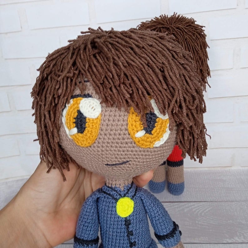 K&ouml;nnte beinhalten: Eine handgefertigte H&auml;kelpuppe mit braunem Garnhaar und gro&szlig;en gelb-wei&szlig;en Augen. Die Puppe tr&auml;gt ein blaues Outfit mit einem gelben Anh&auml;nger. Eine zweite Puppe ist im Hintergrund teilweise sichtbar.