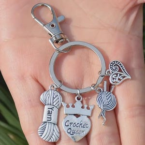 Op de afbeelding: Zilverkleurige sleutelhanger met meerdere bedels. Bedels zijn onder meer een garenspoel met de tekst "Yarn", een hartvormige bedel met de tekst "Crochet Queen", een bol garen met naalden en een decoratief hart. De sleutelhanger heeft een sluiting en een ring.