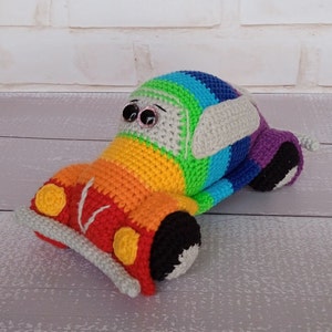 K&ouml;nnte beinhalten: Ein handgefertigtes, geh&auml;keltes Spielzeugauto in Regenbogenfarben. Das Auto hat eine rote Front mit gelben Scheinwerfern, einen grauen Sto&szlig;f&auml;nger und schwarze R&auml;der. Der K&ouml;rper wechselt durch Orange, Gelb, Gr&uuml;n, Blau und Lila. Es hat gro&szlig;e Wackelaugen und graue, ohrenartige Akzente.