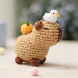 Könnte beinhalten: Handgehäkelte Capybara-Figur in Braun, mit einem kleinen weißen Vogel auf dem Kopf und einer gelben Frucht auf dem Rücken. Das Amigurumi hat braune Akzente und feine Details.