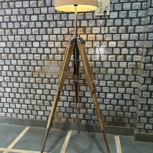 Puede incluir: Una l&aacute;mpara de pie de madera con un tr&iacute;pode y una pantalla de tela beige. La l&aacute;mpara est&aacute; encendida y la luz brilla.