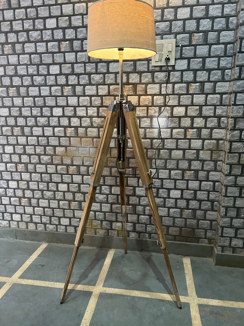 Puede incluir: Una l&aacute;mpara de pie de madera con un tr&iacute;pode y una pantalla de tela beige. La l&aacute;mpara est&aacute; encendida y la luz brilla.