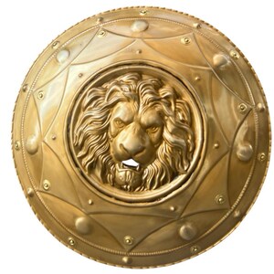 May include: A gold-coloured, round shield featuring a detailed lion's head at the centre. The shield has an embossed design with geometric shapes and rivets around the edge. The lion's face is intricately sculpted, showcasing its mane and facial features.