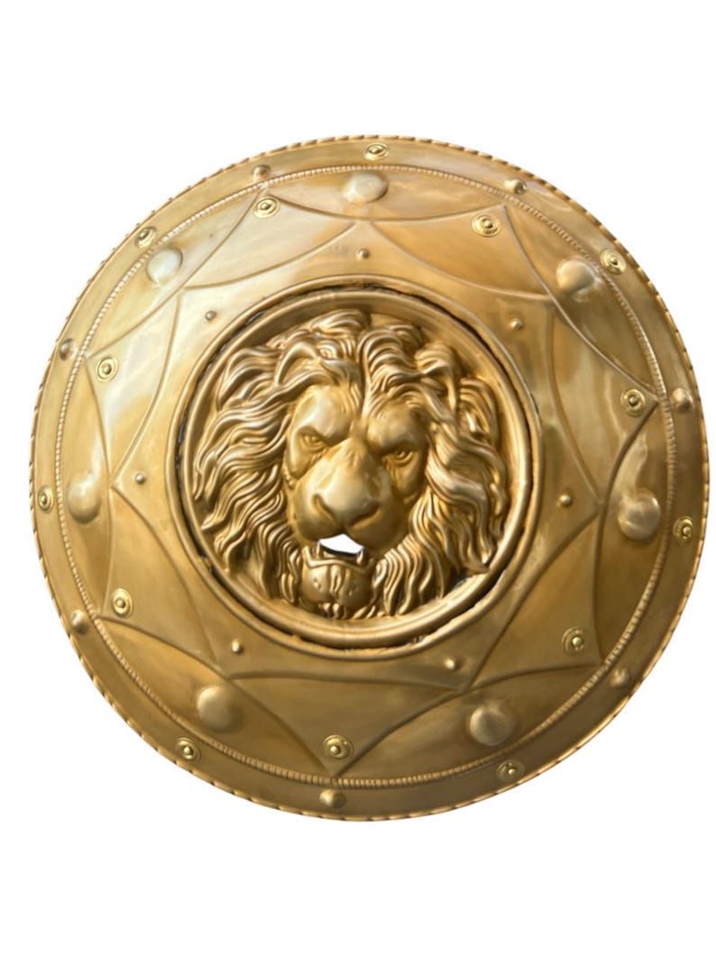 May include: A gold-coloured, round shield featuring a detailed lion's head at the centre. The shield has an embossed design with geometric shapes and rivets around the edge. The lion's face is intricately sculpted, showcasing its mane and facial features.