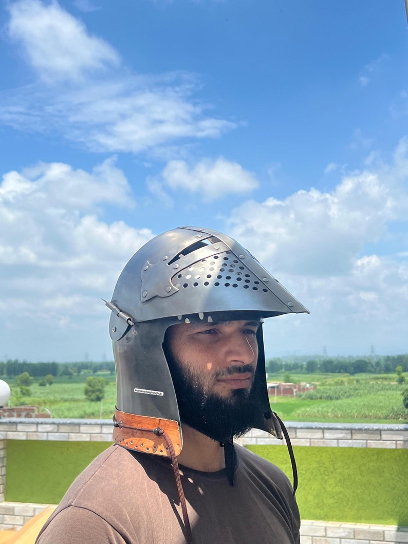 Pode incluir: Um capacete medieval com viseira e uma correia de couro. O capacete &eacute; feito de metal e tem uma superf&iacute;cie texturizada. O capacete &eacute; usado por uma pessoa com barba.