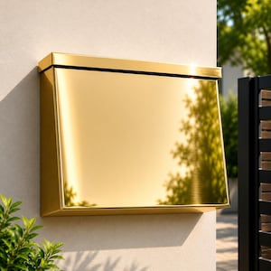 May include: A rectangular wall-mounted mailbox with a highly polished, reflective gold-tone metallic finish. The mailbox features a flat top lid and a smooth, angled front panel that mirrors the surrounding environment, mounted on a light wall.