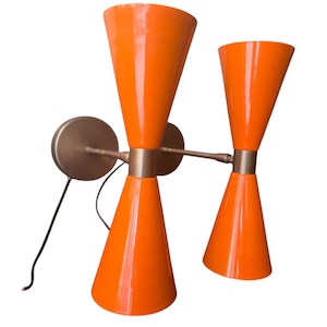 Può includere: Un paio di applique da parete arancioni con accenti in ottone. Le applique hanno un design a forma di cono e sono montate su una parete.