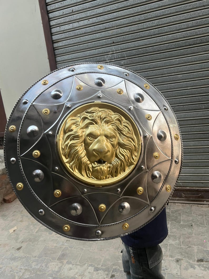 May include: A circular shield in silver with a gold lion's head emblem at its centre. The shield has a decorative border with rivets and circular accents. The lion's head is detailed with a mane and facial features. The shield appears to be made of metal.