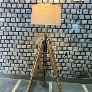 Puede incluir: Una l&aacute;mpara de pie de tr&iacute;pode de madera con una pantalla de tela beige. La l&aacute;mpara est&aacute; enchufada a una toma de corriente en una pared de ladrillo.