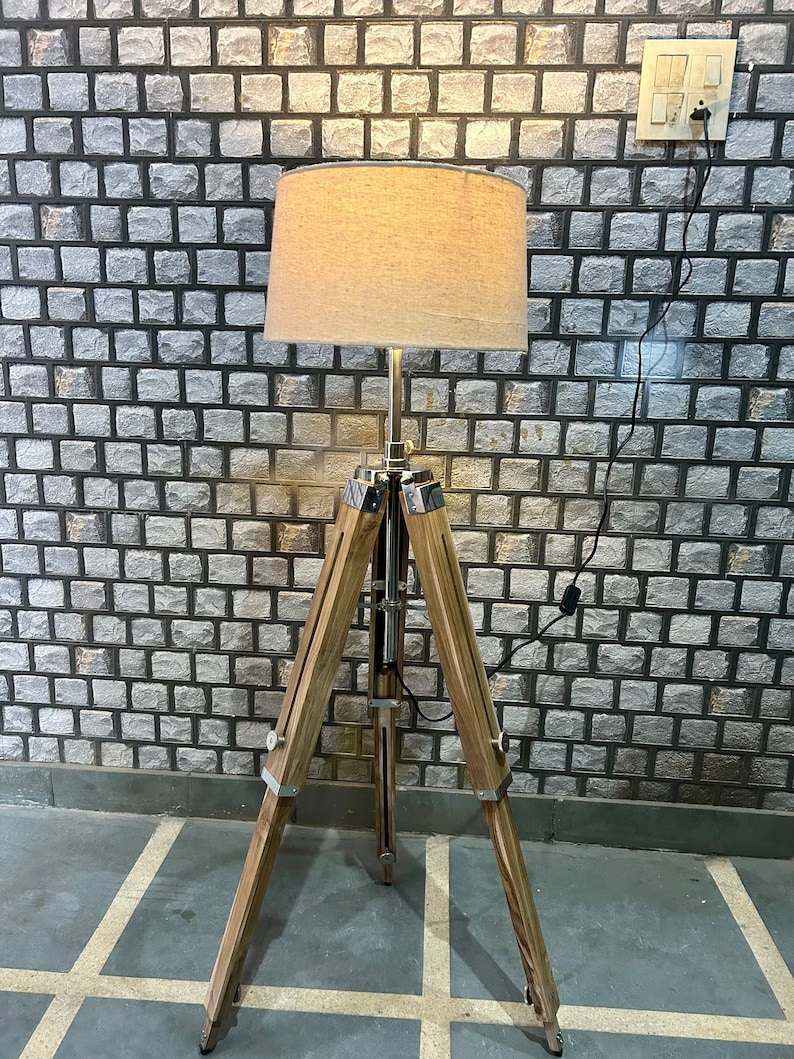 Puede incluir: Una l&aacute;mpara de pie de tr&iacute;pode de madera con una pantalla de tela beige. La l&aacute;mpara est&aacute; enchufada a una toma de corriente en una pared de ladrillo.