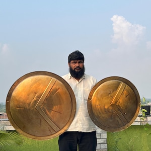 May include: Two round, gold-colored shields with a Greek key design. The shields are held by a person in front of a brick wall.