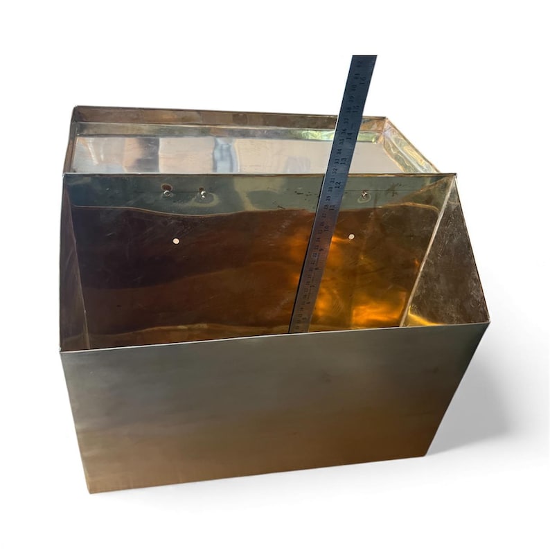 May include: A rectangular brass wall-mounted bin with an open top and a hinged lid. A metal ruler is placed inside, showing a height measurement of approximately 12 inches. The brass has a polished, reflective surface with visible mounting holes.
