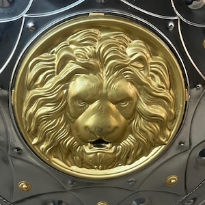May include: Close-up of a medieval shield featuring a gold lion's head emblem. The shield has a silver-coloured metal base with gold rivets and decorative elements. The lion's head is a detailed, golden relief.