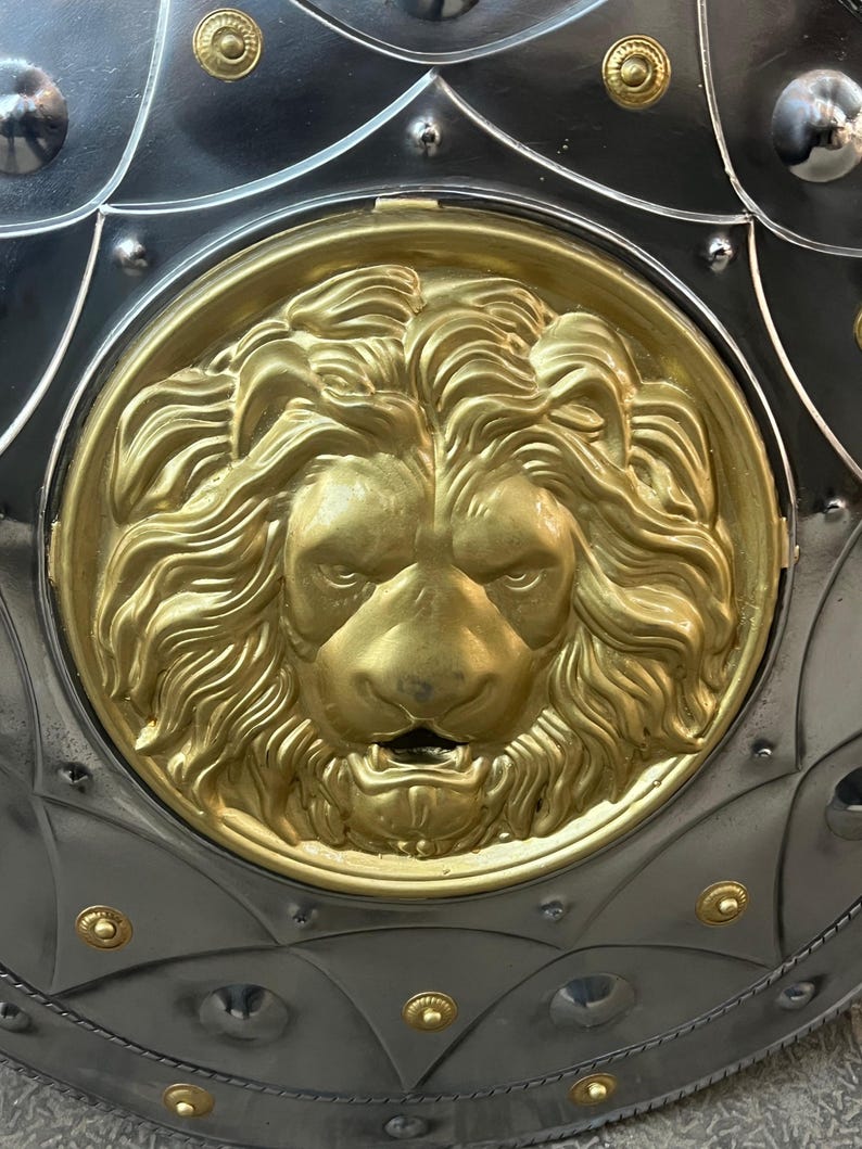 May include: Close-up of a medieval shield featuring a gold lion's head emblem. The shield has a silver-coloured metal base with gold rivets and decorative elements. The lion's head is a detailed, golden relief.