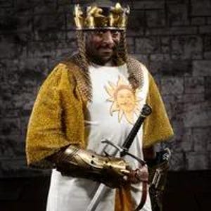 May include: A man wearing a gold crown and chainmail armor, with a white tunic featuring a gold sun design. He is holding a sword in his right hand.