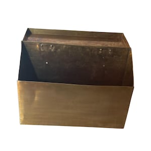 May include: A rectangular brass wall-mounted mailbox with a hinged lid. The metal surface has a warm, metallic gold tone with visible signs of age and patina. The structure features a flat front panel and a top cover attached with metal rivets.