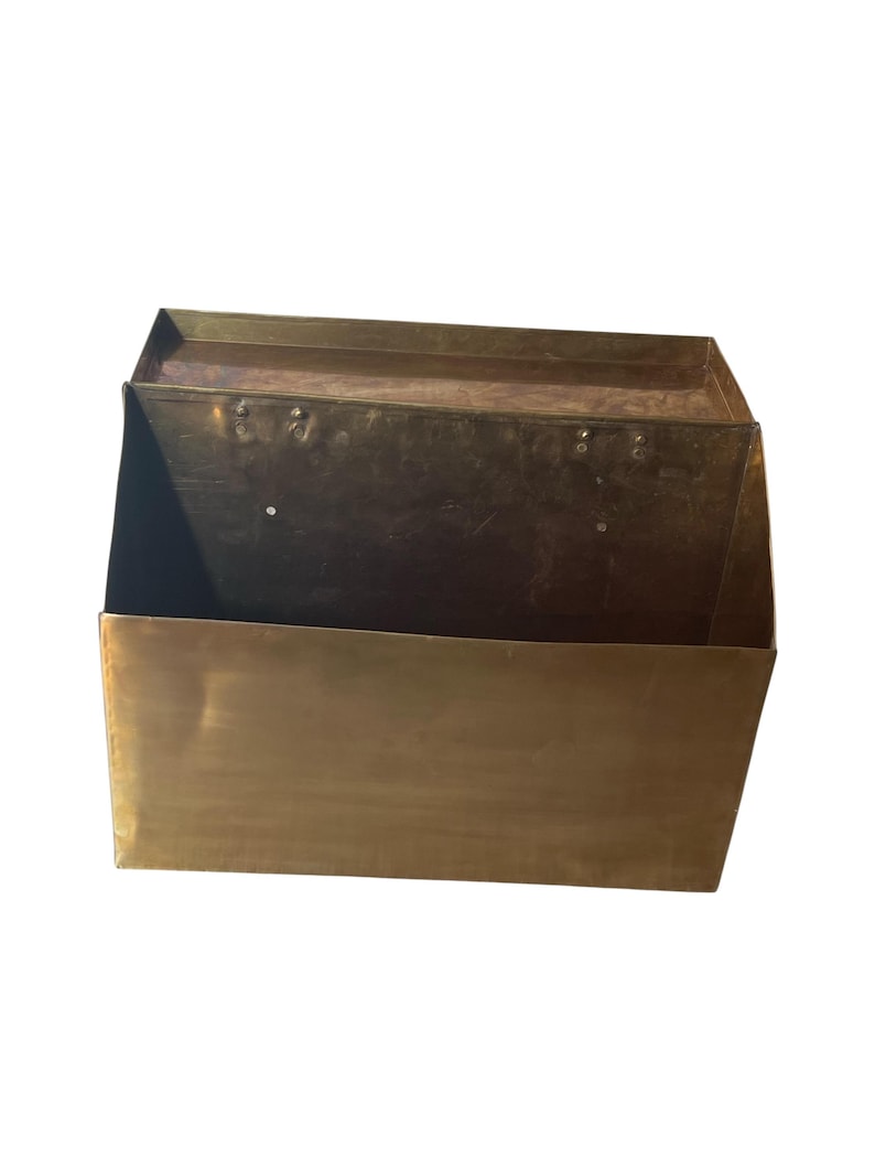 May include: A rectangular brass wall-mounted mailbox with a hinged lid. The metal surface has a warm, metallic gold tone with visible signs of age and patina. The structure features a flat front panel and a top cover attached with metal rivets.