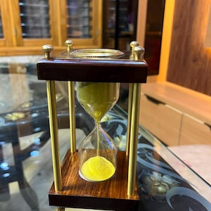 Puede incluir: Un reloj de arena con marco de madera marrón oscuro, cuatro pilares de latón pulido y una incrustación circular de latón en la parte superior. El bulbo de vidrio transparente contiene arena amarilla brillante fluyendo hacia abajo.