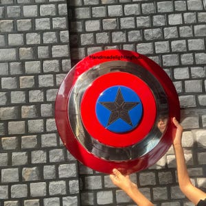 Puede incluir: Un escudo rojo, plateado y azul con un emblema de estrella. El escudo tiene un dise&ntilde;o circular con un anillo exterior rojo, un anillo central plateado y un centro azul con una estrella negra. El escudo est&aacute; sostenido por una persona.