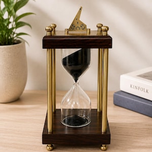 Peut inclure: Un sablier avec un cadre en bois foncé, quatre piliers en laiton et un ornement supérieur en laiton. Le bulbe en verre contient du sable noir, dont une partie se trouve dans la chambre supérieure. La base et le sommet sont carrés avec des pieds.