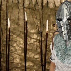 Pode incluir: Um cavaleiro com um capacete prateado e um colar de malha est&aacute; em frente a uma parede com v&aacute;rias estacas de madeira.
