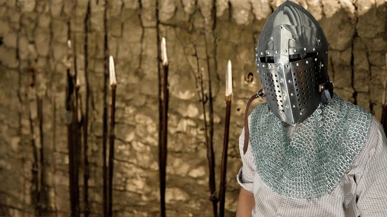 Pode incluir: Um cavaleiro com um capacete prateado e um colar de malha est&aacute; em frente a uma parede com v&aacute;rias estacas de madeira.