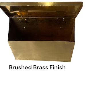May include: A rectangular metal wall-mounted mailbox with a hinged lid and an open top section. The object features a metallic gold-toned surface with a brushed brass finish. The text "Brushed Brass Finish" is printed below the object.