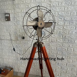 Op de afbeelding: Een vloerventilator met een metalen kooi en vier bladen, voorzien van een centraal embleem met de tekst Made for Royal Navy London. De ventilator is gemonteerd op een houten statief met drie poten, metalen accenten en een zwart snoer met schakelaar.