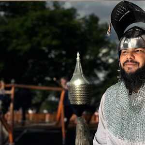 Pode incluir: Um homem vestindo um capacete prateado com um colar de malha e uma camisa branca. Ele tem barba e est&aacute; olhando para a c&acirc;mera.