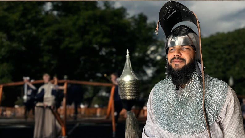 Pode incluir: Um homem vestindo um capacete prateado com um colar de malha e uma camisa branca. Ele tem barba e est&aacute; olhando para a c&acirc;mera.