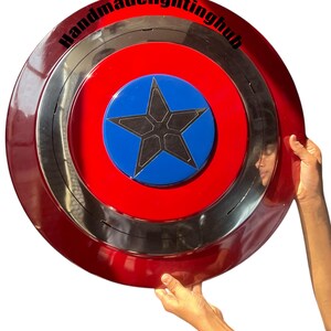 Puede incluir: Un escudo rojo, plateado y azul con un dise&ntilde;o de estrella. El escudo tiene forma circular, con un anillo exterior rojo, un anillo central plateado y un centro azul con una estrella negra. El texto "Handmadelightinghub" est&aacute; impreso en el anillo exterior.